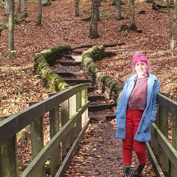 A photo of Bryony standing on a bridge in an autumn woodland. Her pink hair is tied up, she wears a long blue coat and red trousers. The bridge and the forest floor behind her are covered in fallen leaves.