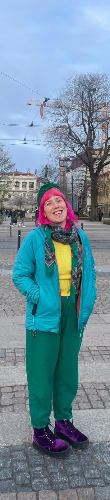 Bryony is standing in a city square. She is wearing a blue coat, a yellow jumper, green trousers and purple velvet boots. She is smiling to the camera, her head tilted to one side.