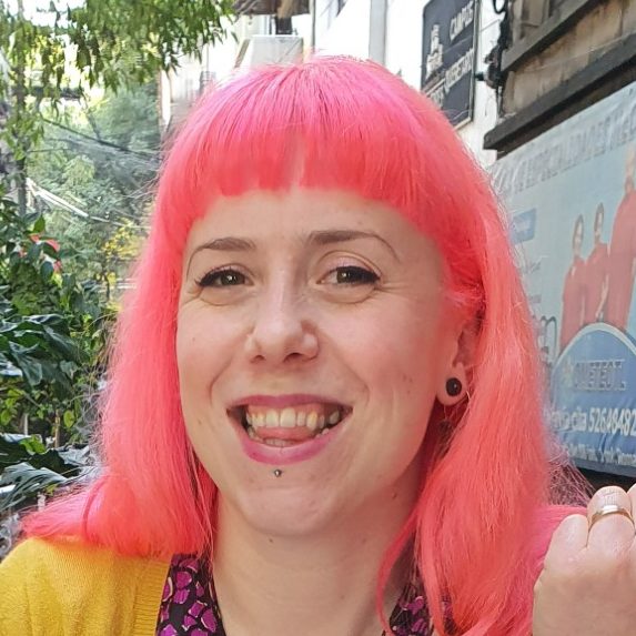 A photo of Bryony. She has bright, coral coloured hair. She has a fringe, brown eyes and a lip piercing. She is looking towards the camera and smiling.