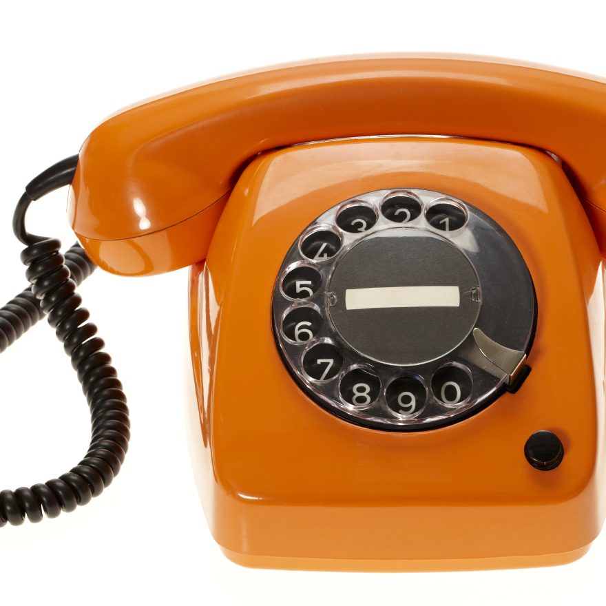 A photo of a bright orange vintage telephone with a round black dialpad and a curly black cord.